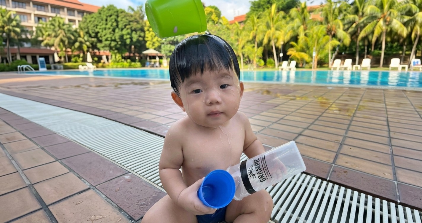 A toddler at the poolside, mid water-play, looking curiously at the camera